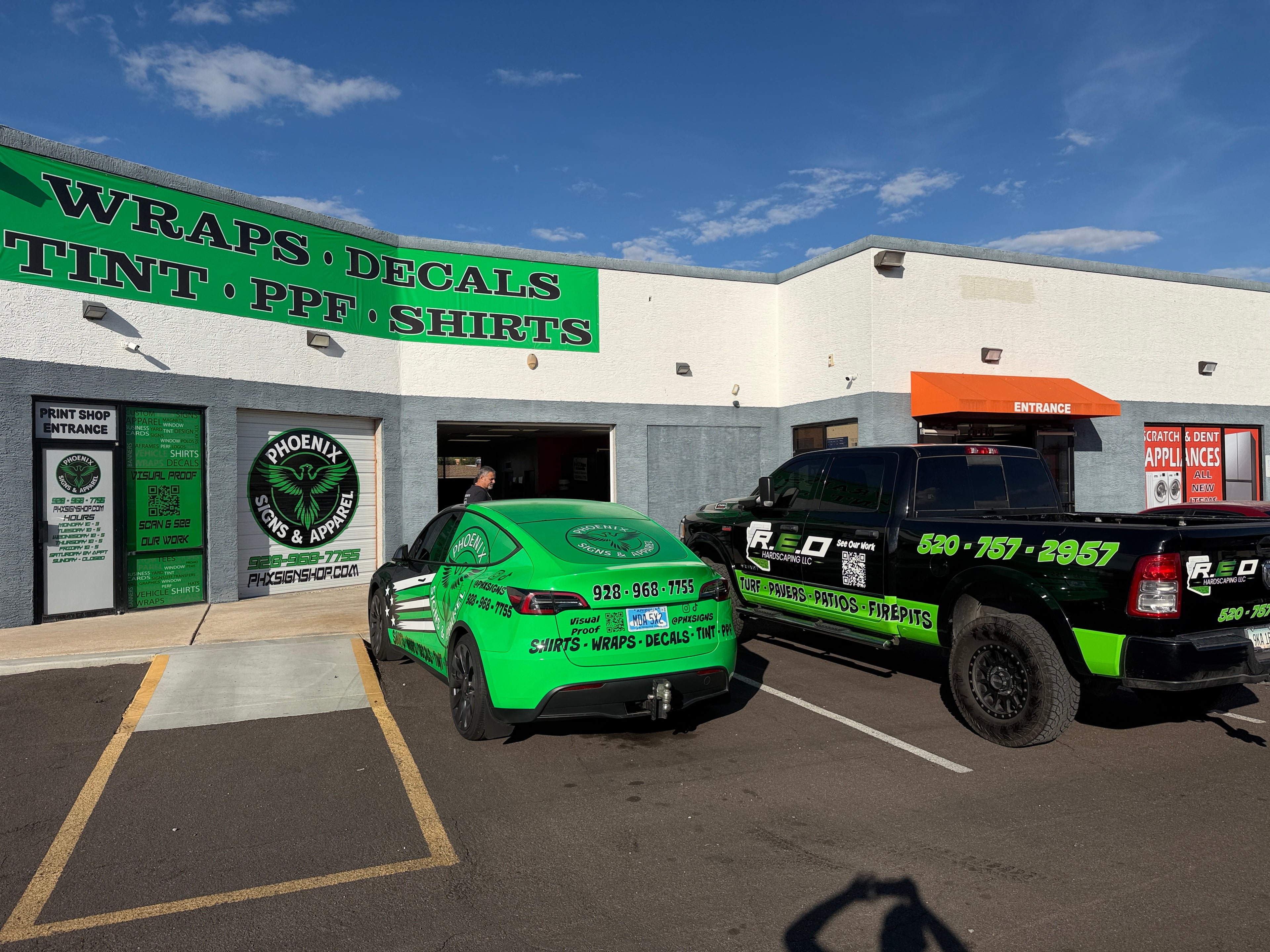 Green car and black truck in front of a business with signs for vehicle wraps, decals, and PPF.