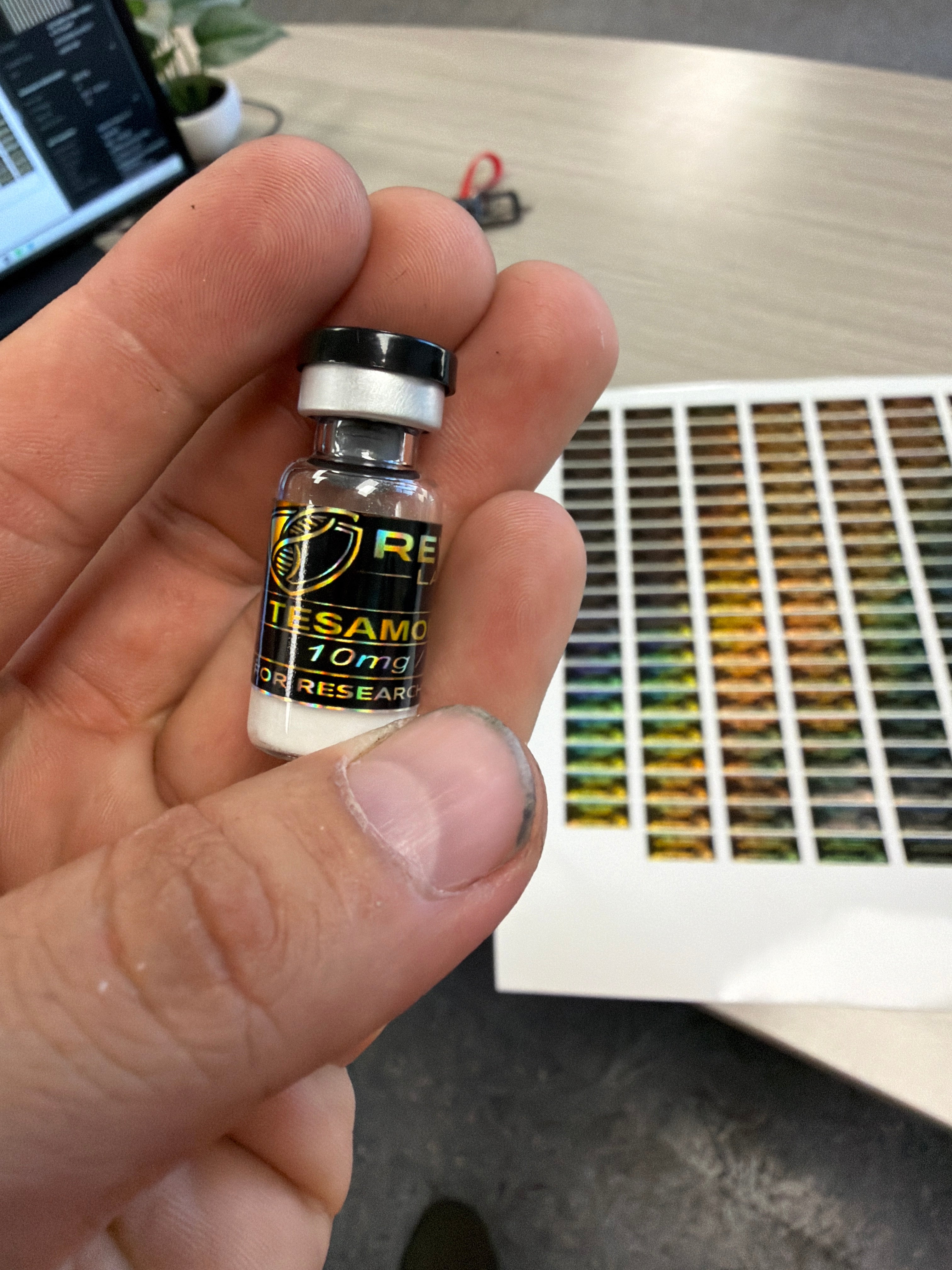 A hand holding a small glass vial labeled with new holographic labels made by Phoenix Signs & Apparel. Printed label sheet is behind the hand.
