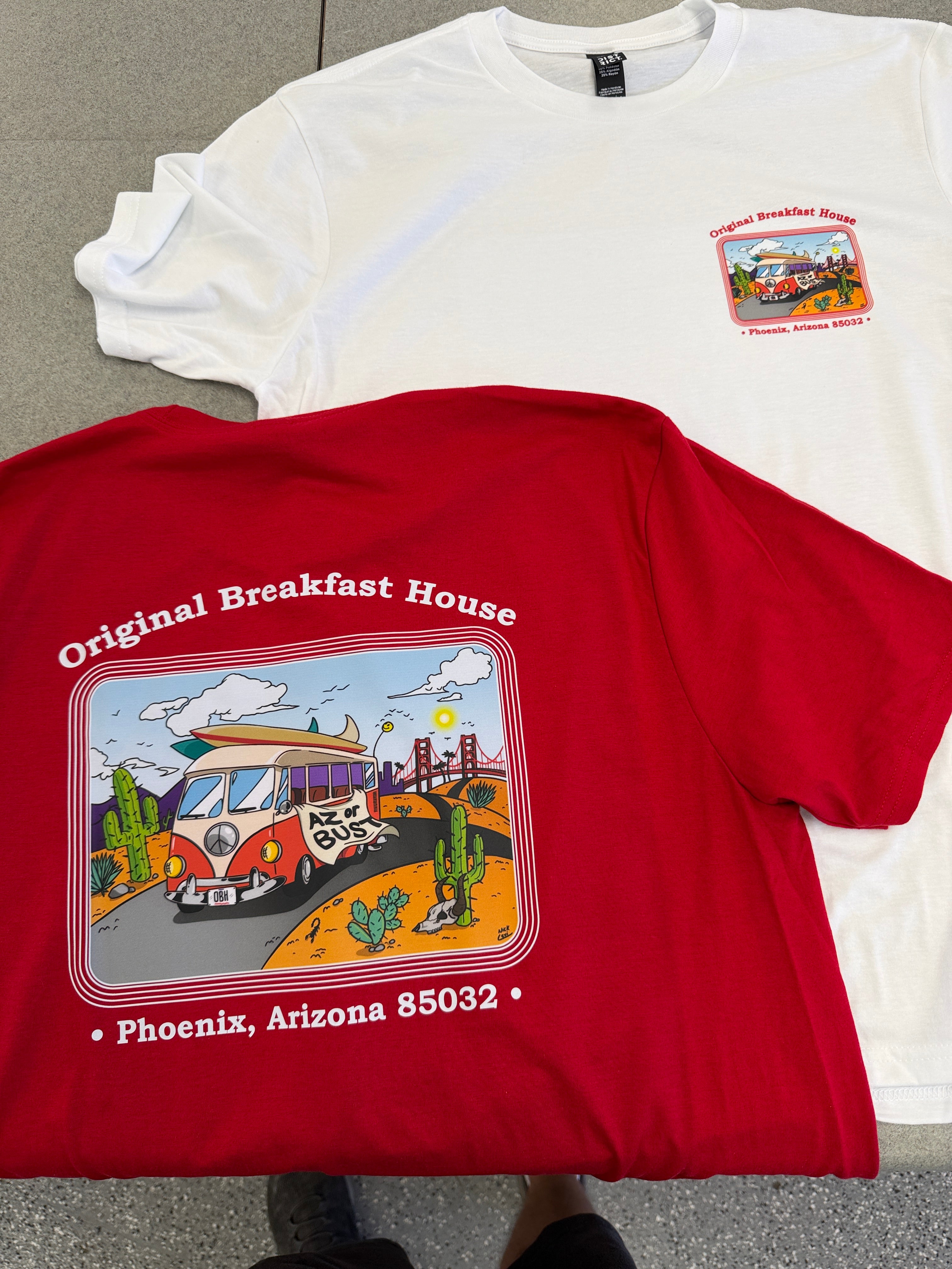 Two graphic t-shirts laid flat on a gray floor: a white short-sleeve shirt with a small multicolor patch graphic on the chest, and a red long-sleeve shirt with a large colorful 'Original Breakfast House' restaurant graphic on the front reading 'Phoenix, Arizona 85032'.
